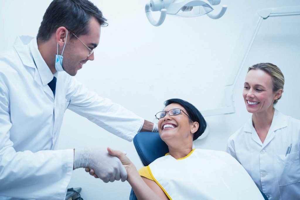 Male dentist shaking hands with female patient in chair. Dental hygienist by their sides, everyone is smiling.