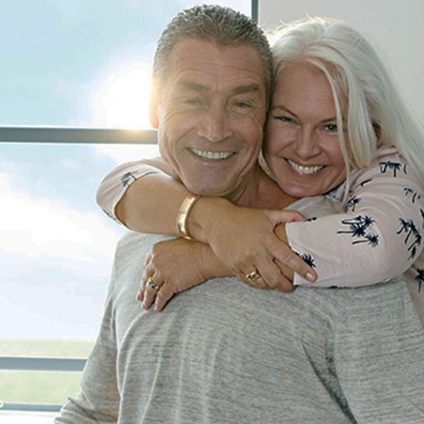 Middle aged couple in kitchen, woman hugging man from behind, both smiling at the camera