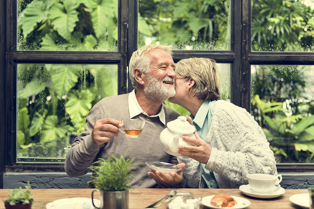 Older couple enjoying hot tea and pastries during a rainy day, and the wife is whispering in her husband’s ear