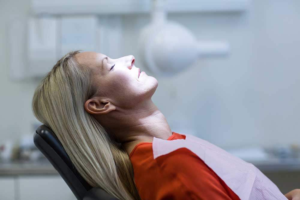 Woman laying in a dentist chair with her eyes shut and the light shining on her as she prepares for treatment