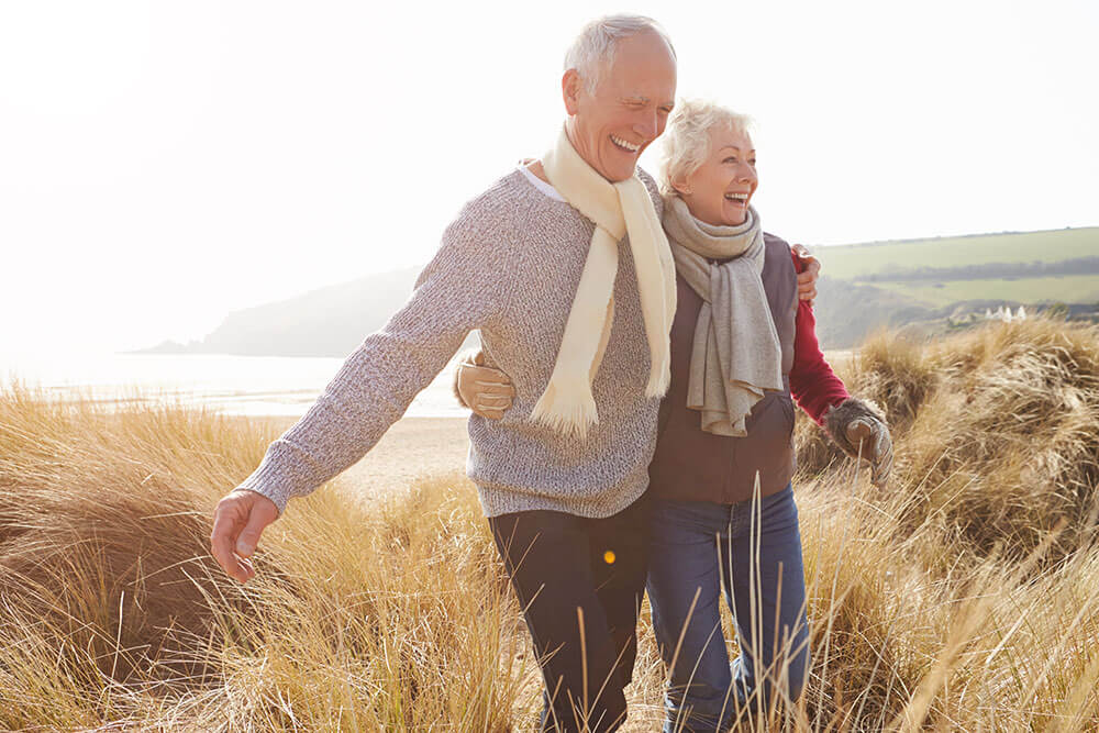 Smiling older couple walking arm in arm through the grass with green hills and a beach in the background